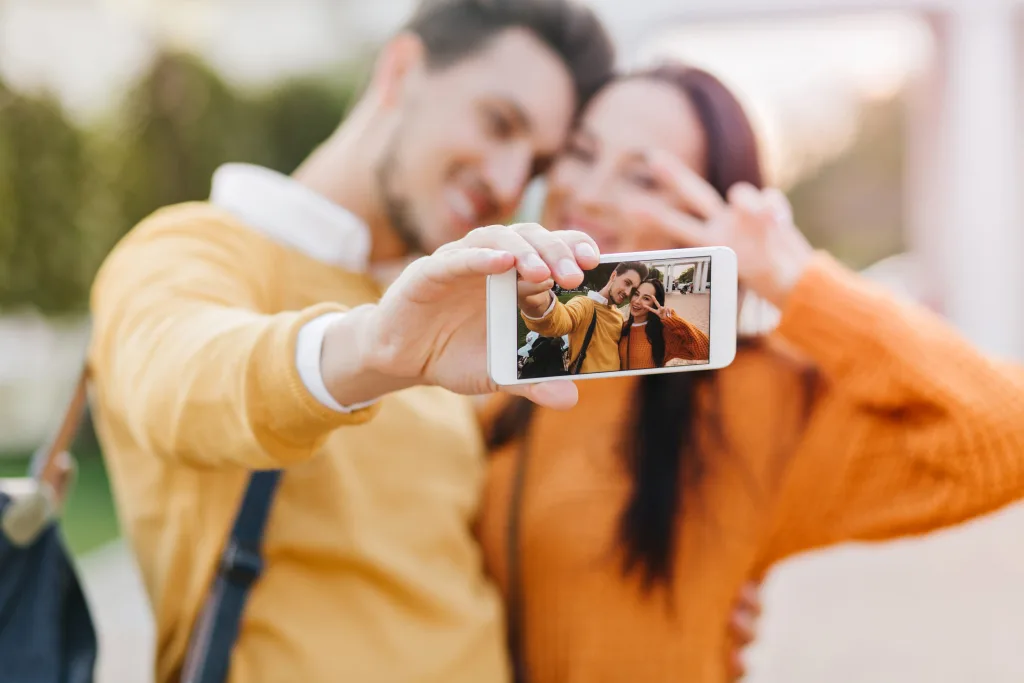 Фото о любви.
Cute girl posing with peace sign while her boyfriend in orange sweater making selfie. Funny couple using smartphone for outdoor photoshoot in autumn day.